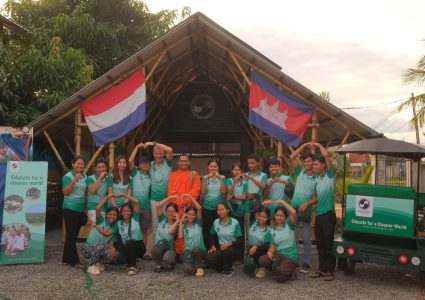 Group of young Cambodian students and the co-founders in green shirts with Chikara Cambodia logo, and a monk in orange monk clothing, in front of a bamboo building. On the left a green banner and on the right a green tuk-tuk.