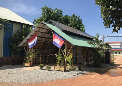 Bamboo building with light green roof and in front a Dutch flag and a Cambodian flag.