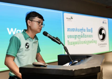 Jonge Cambodjaanse man in Chikara Cambodia poloshirt spreekt in een microfoon achter een katheder tijdens Waste Summit Cambodia 2026, met Khmer-tekst op schermen op de achtergrond.